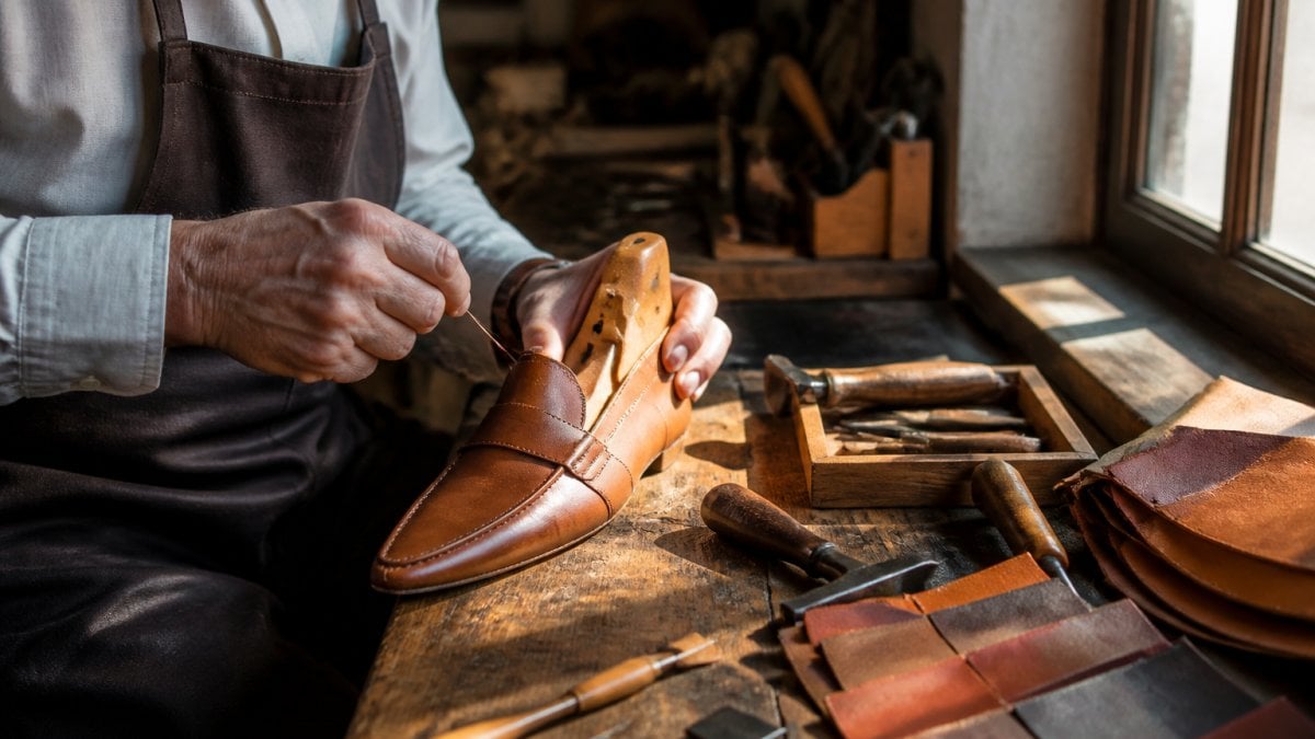 Close-up of a craftsman's hands stitching a calf leather loafer on a wooden last in a Spanish workshop, hand tools and leather swatches on the bench, warm window light
